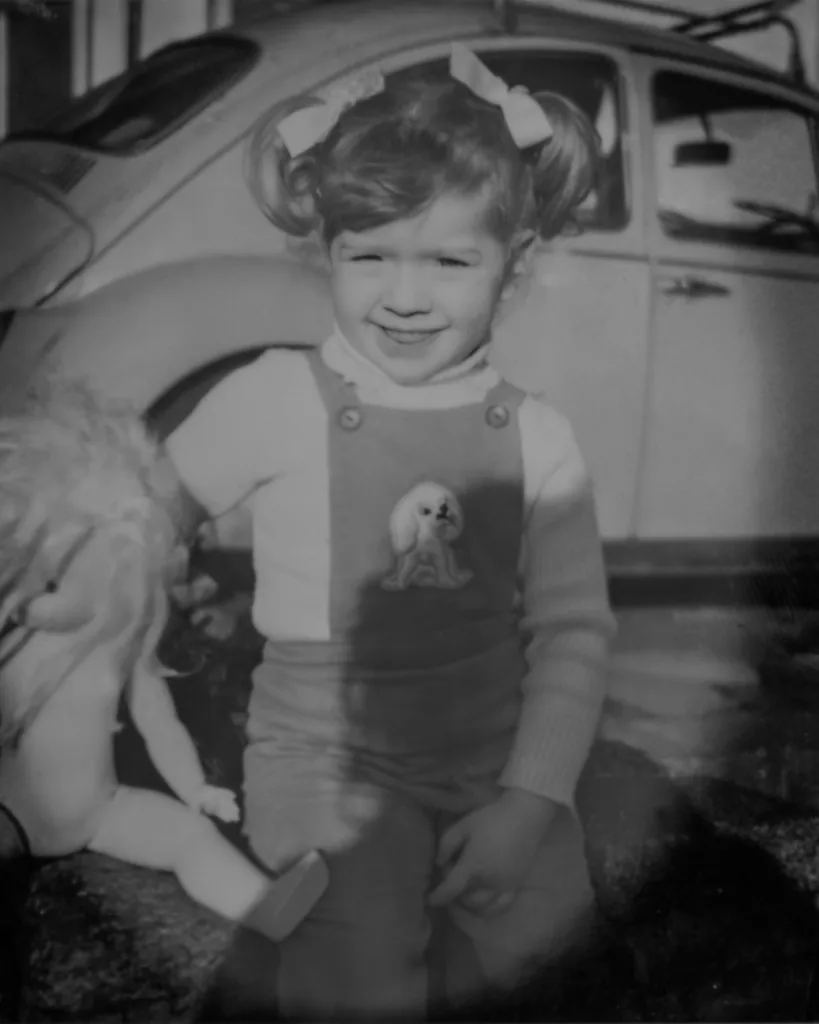 a little girl with pigtails, wearing cord dungarees infront of a VW beetle holding her doll